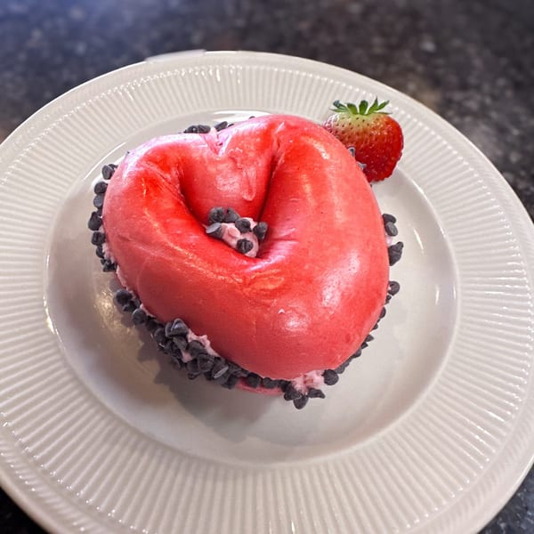 A heart-shaped red bagel with cream cheese and chocolate chips, garnished with a strawberry.