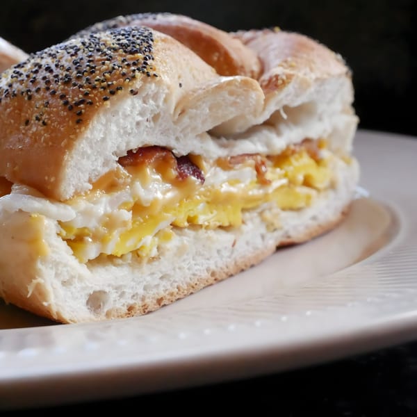Close-up of a poppy seed bagel sandwich with scrambled eggs and bacon on a white plate.