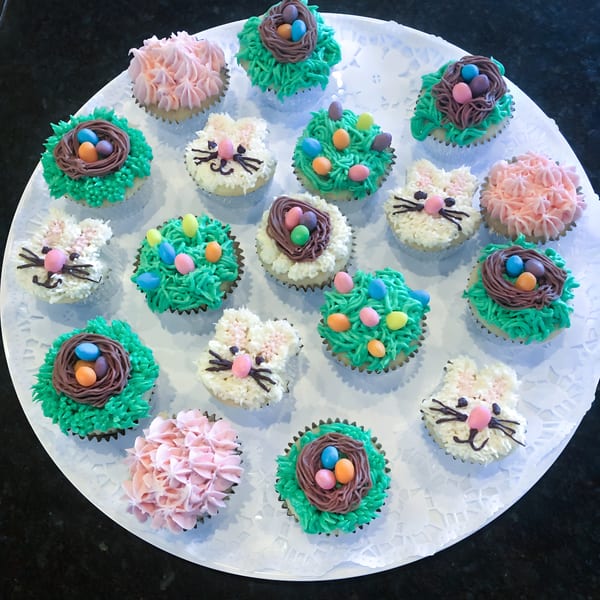 A white platter holds Easter-themed cupcakes, some with bunny faces, others with nests and candy eggs.